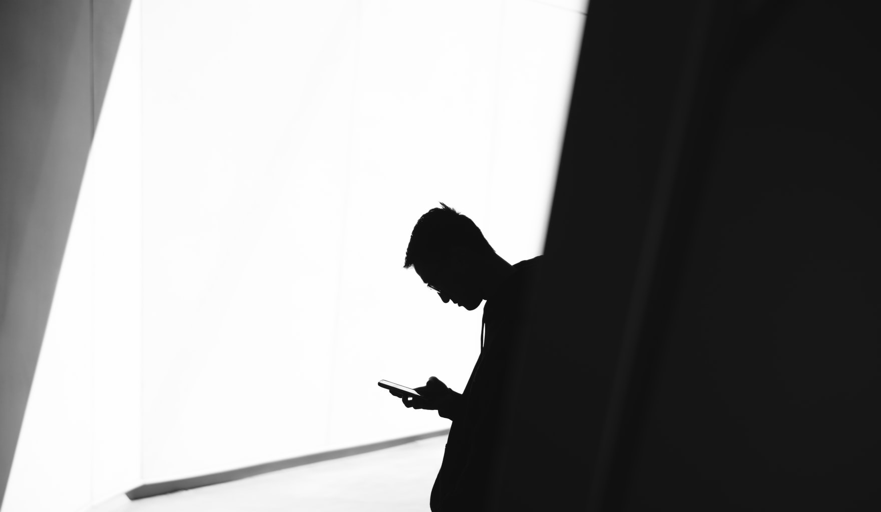 A silhouete of a man leaning against a wall looking at his phone.